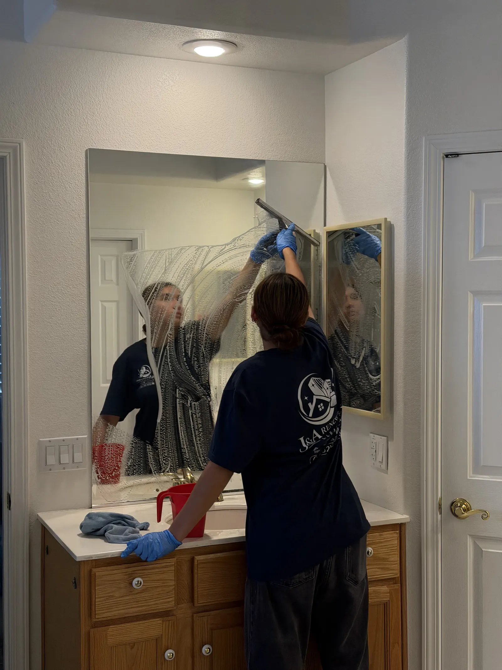 Cleaning a bathroom mirror with a squeegee, in a Renew & Shine shirt
