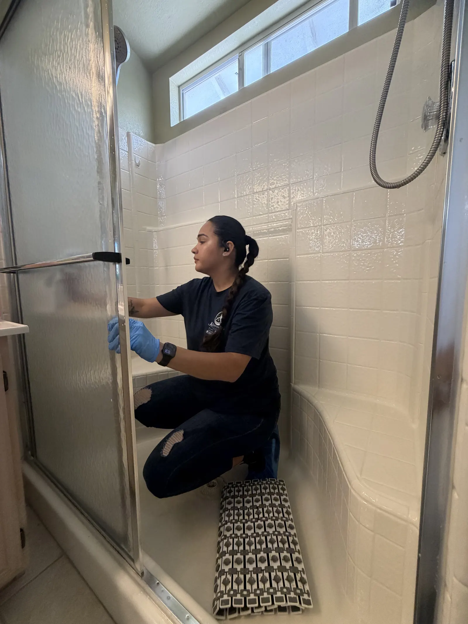Ambar, in blue gloves, cleaning the glass door of a tiled shower